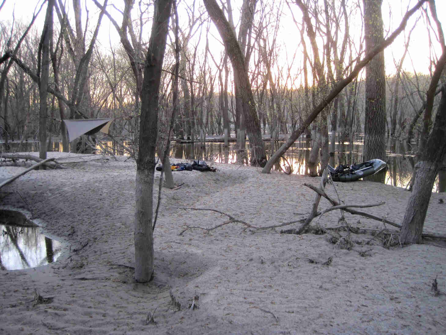 A makeshift camping area on a sandbar with trees, a tarp tent and an inflatable raft, in a flooded river bottoms area